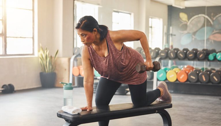 Woman in her 40s performing a dumbbell row on a bench in a real gym β strength training to reduce menopause belly fat